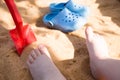 baby& x27;s legs in the beach sand, blue flip flops and a toy shovel, play in the sandbox