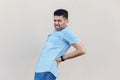 Back ache or kidney pain. Portrait of sick handsome young bearded man in blue shirt standing and holding his painful back