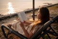 Brunette reading book while resting on deck chair at sunset Royalty Free Stock Photo