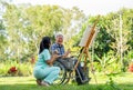 Back of Asian nurse woman sit near senior man on wheelchair enjoy to paint the painting outdoor in the garden and they look Royalty Free Stock Photo