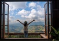 Back of Asian woman with freedom arms open standing behind large wooden window with mountain view and cloudy blue sky at Doi Chang