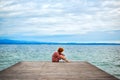 Back of faceless red haired woman in red dress sitting on wooden pier on Garda lake in cloudy day. Italian vacation. Garda lake Royalty Free Stock Photo