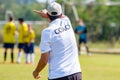 Back of football coach wearing white COACH shirt at an outdoor sport field coaching his team during a game