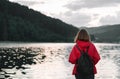 The back of a girl in a red raincoat on the background of a mountain lake and forest, carrying a backpack. Girl tourist stands in Royalty Free Stock Photo