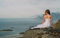 Back of girl in wedding dress sitting on rocks in mountains and looking to fjord