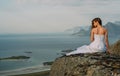 Back of girl in wedding dress sitting on rocks in mountains and looking to fjord