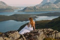 Back of girl in wedding dress sitting on rocks in mountains and looking to fjord