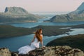 Back of girl in wedding dress sitting on rocks in mountains and looking to fjord