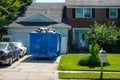 The back of a large blue dumpster in the driveway in front of the garage of a house on a sunny day