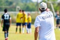 Back of male football coach wearing white COACH shirt at an outdoor sport field coaching his team during a game