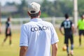 Back of male football coach wearing white COACH shirt at an outdoor sport field coaching his team during a game