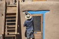 Back of Native American man in shorts and tee-shirt and sun hat painting bright blue-turquoise around the door of a mud adobe