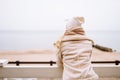 Back shot of blonde haired woman enjoy seaview at winter day. Female wearing a light jacket, hat, scarf and have hot