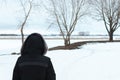 A back shot of a woman wearing a warm fur coat and a hood. Landscape shot in the background, leafless branches of trees and a