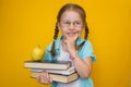 Back to school. Beautiful little girl with pigtails and glasses pensively smiles and holds textbooks and a yellow apple on a Royalty Free Stock Photo