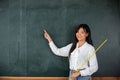 Asian female teacher smiling with wooden stick pointing to blackboard