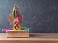 Back to school concept with rocket, pencils, notebook and book on wooden table over blackboard background Royalty Free Stock Photo