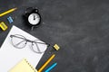 Back to school concept. School stationery, glasses, paper notebook and clock on black board background. Flat lay, top view,