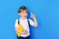 Back to school concept. Schoolboy boy with backpack and holding yellow book in front of blue background. Child shows Royalty Free Stock Photo