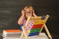 Back to school. A funny little girl in round glasses is sitting at a table against the background of a blackboard. Royalty Free Stock Photo