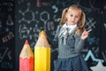 Back to school! A little blonde girl in school uniform stands with two very large red and yellow pencils on the floore against