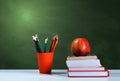 Back to school, orange pencil holder, stack of books on white table with red apple, empty green school board background, education Royalty Free Stock Photo