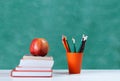 Back to school, orange pencil holder, stack of books on white table with red apple, empty green school board background, education Royalty Free Stock Photo