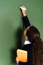 back view of african american schoolgirl writing on chalkboard while holding book. Royalty Free Stock Photo