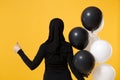 Back view Arabian muslim woman in hijab celebrating holding black white air balloons isolated on yellow background