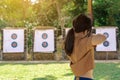 Back view of Asian girl wear face mask aims archery bow and arrow to colorful target in shooting range during training and