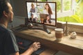 Back view of Asian man in casual wear talking to his parrtner about work plant in video conference by computer on wooden table