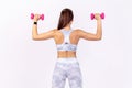 Back view athletic woman in white sportswear raising hands with pink dumbbells, developing strength and power, pumping up muscles