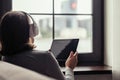 Back view of ÃÂaucasian young woman with tablet in headphones listening music sitting near window of apartments. Royalty Free Stock Photo