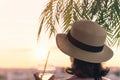 Back view of a beautiful girl with cold water in a glass in a straw hat against the background of the sea in branches of palm tree Royalty Free Stock Photo