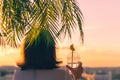 Back view of a beautiful girl with lemon water in a plastic glass against the background of the sea in branches of palm trees.