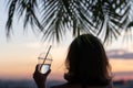 Back view of a beautiful girl with mineral water in a glass against the background of the sea in branches of palm trees. Sunset