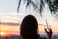 Back view of a beautiful girl with mineral water in a glass against the background of the sea in branches of palm trees. Sunset