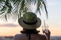 Back view of a beautiful girl with mineral water in a glass in a straw hat against the background of the sea in branches of palm