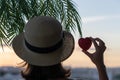 Back view of a beautiful girl in a straw hat holding red heart against the background of the sea in branches of palm trees. Sunset Royalty Free Stock Photo