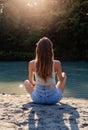 Back view of beautiful young woman with long hair sitting alone on sand looking at river and sun Royalty Free Stock Photo