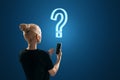 Back view of blonde young casual woman with smartphone looking for answers. Glowing question mark hologram above head on blue