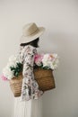 Back view of boho girl holding pink and white peonies in rustic basket. Stylish hipster woman in hat and bohemian floral dress