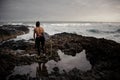 Back view brunette boy standing in the black swimsuit with naked torso with a white surf in his hands on the shore Royalty Free Stock Photo