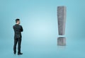 Back view of a businessman looking at big 3D concrete exclamation mark isolated on blue background