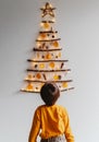 Back view of child looking at handmade craft Christmas tree made from sticks and natural materials hanging on wall.