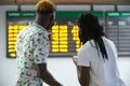 Back view of couple in a train station wearing masks, looking at their smartphone and the travel board Royalty Free Stock Photo