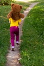 Back view. curly smiling girl in a yellow T-shirt holds a large brown teddy bear on her neck and runs along the path