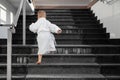 Back view of cute funny caucasian blond kid boy wearing white pool spa bathrobe walking upstairs on big marble staircase