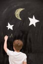 Back view on cute kid boy drawing stars with chalk on a black board with white stars and moon. Child writing on wall