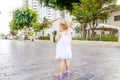 Back view cute little blondy toddler girl in white dress standing along on the alley in the city park. Childhood concept. Soft sel